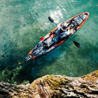 A person paddling a full-sized recreational kayak on clear turquoise water, viewed from an overhead cliffside perspective.